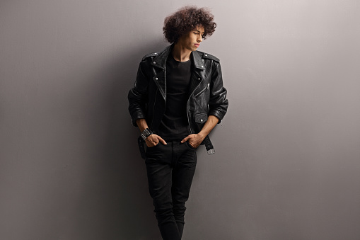 Young man with a leather jacket and curly hair leaning on a gray wall