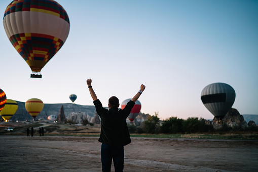 Back view of man with arms raised up watching colorful hot air balloons landing in mountains excited with achievement and life success