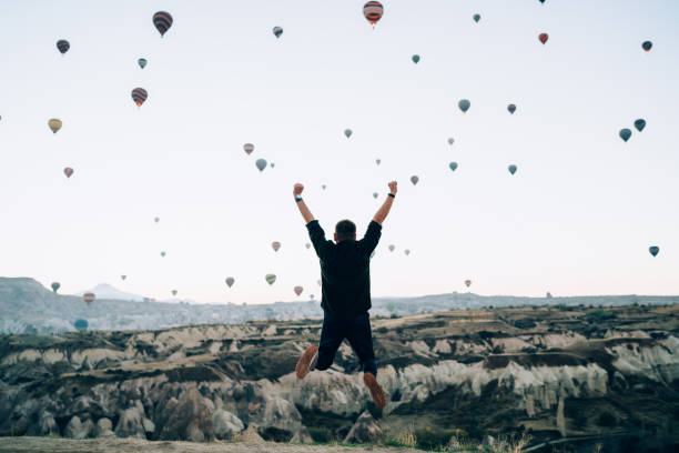 Excited man jumping while raising hands up against hot air balloons Aerial back view of male winner raising hand up while jumping against hot air balloons landing in mountain at sunrise jump jet stock pictures, royalty-free photos & images