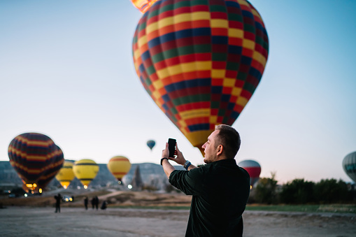 Back view of casual traveling man in black shirt taking photo of flying air balloons with smartphone standing in mountains