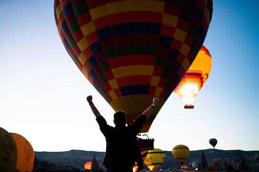 Happy casual man from behind raising arms in air and celebrating success while looking at flying colorful hot air balloons above mountains