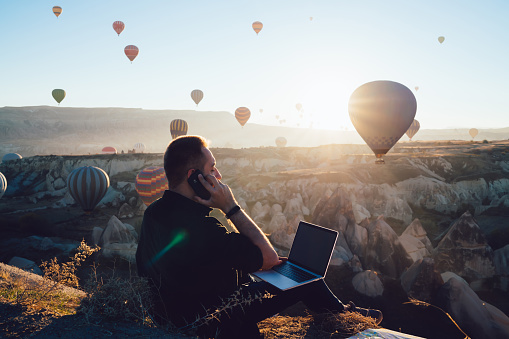 Side view of male traveler browsing modern laptop and answering phone call while sitting near rocky terrain and hot air balloons in back lit