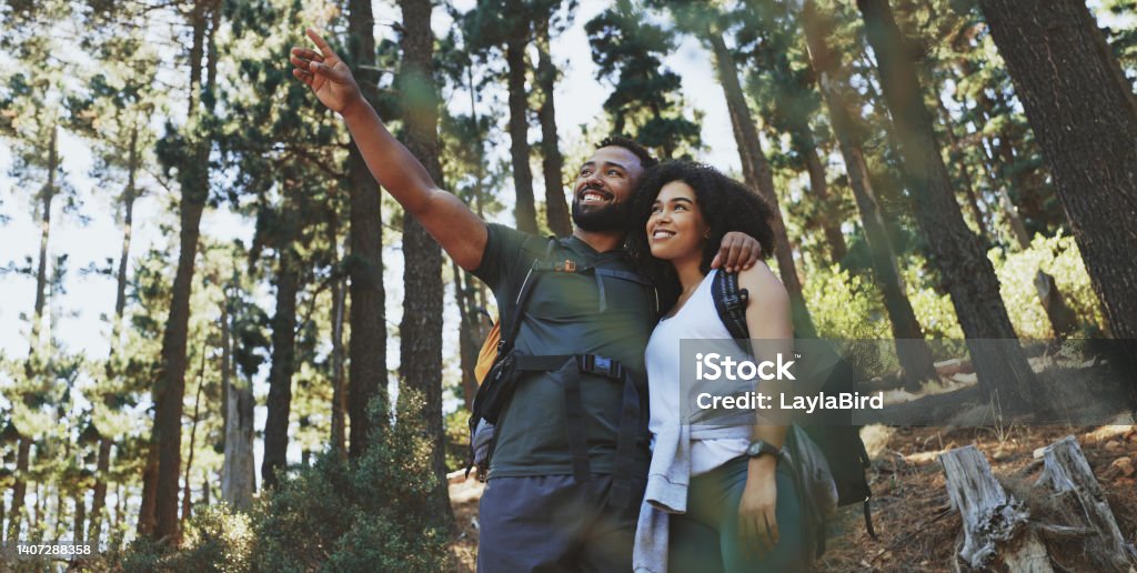 Happy couple hiking a trail and enjoying the view in a forest. Cheerful adventurous man and woman pointing at scenery while taking a break from exploring their surroundings during a trek in nature Happy young couple hiking a trail in the forest. Cheerful adventurous man and woman pointing at scenery while taking a break to admire the view and explore their surroundings during a trek in nature African-American Ethnicity Stock Photo