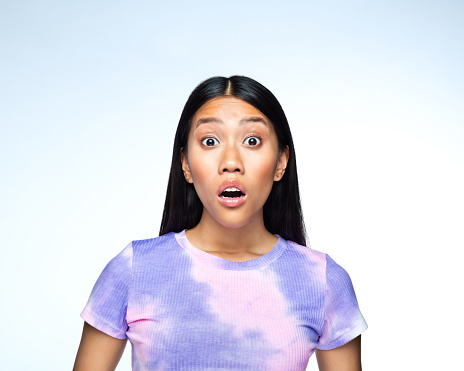 Headshot of surprised young woman wearing lilac tie dye top, looking away with mouth open. Studio shot against blue background.