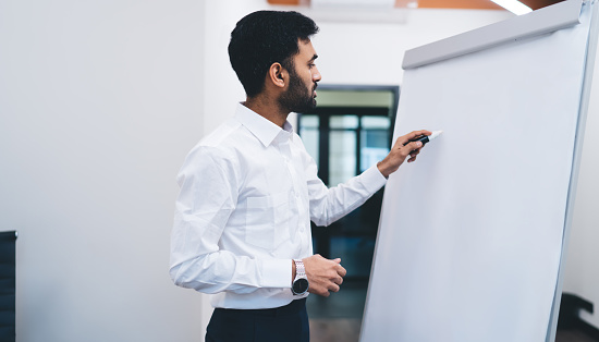 Side view of successful man with marker in hand clarifying financial details on white board while working in large company