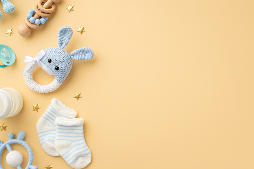 Baby accessories concept. Top view photo of knitted bunny rattle toy blue teether wooden rattle milk bottle tiny socks soother and gold stars on isolated pastel beige background with copyspace