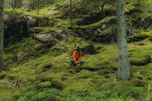 Female traveler in orange enjoying a day in the scenic Swedish forest, sitting on the soft moss and looking at the wonderful nature