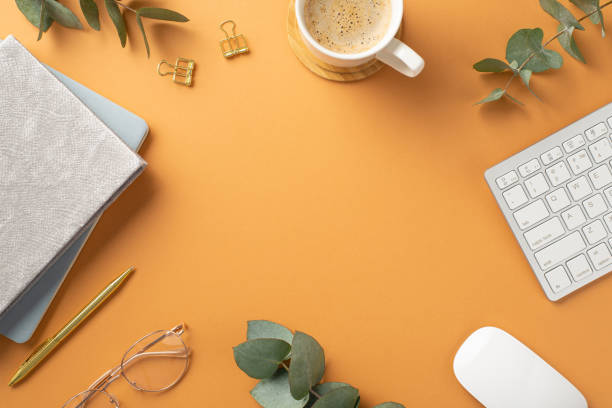 Top view photo of workstation keyboard computer mouse cup of coffee on wooden stand glasses planners gold pen binder clips and eucalyptus on isolated orange background with copyspace in the middle vista superior foto de la estación de trabajo teclado computadora ratón taza de café en vidrios de soporte de madera planificadores de carpetas de plumas de oro clips y eucalipto sobre fondo naranja aislado con espacio de copia en el medio