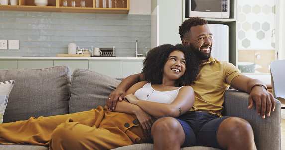 Couple daydreaming and cuddling on couch at home. Husband and wife sitting on sofa. Man embracing his female partner. Girlfriend and boyfriend laughing together. Man and woman thinking and bonding