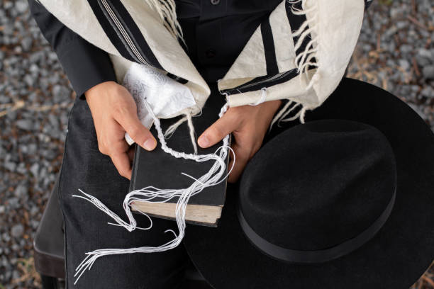 nahaufnahme eines juden, der einen siddur hält, auf dem der tzitzit seines tallit und daneben ein hut ruht. - judaism jewish ethnicity hasidism rabbi stock-fotos und bilder