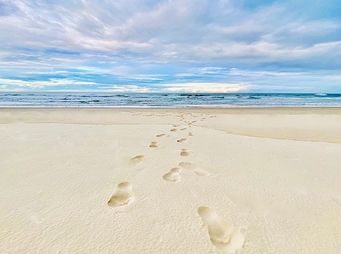 Horizontal close up seascape of barefoot footprints in sand with coastal ocean waves horizon under pastel dusk cloudscape at Brunswick Heads near Byron Bay NSW Australia