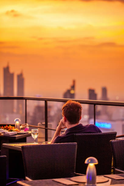 istock Young man with glass of wine rest at luxury rooftop restaurant watching orange sky sunset. Male with cocktail drink at sky bar terrace looking at modern city skyline. Skyscrapers on background 1407299577
