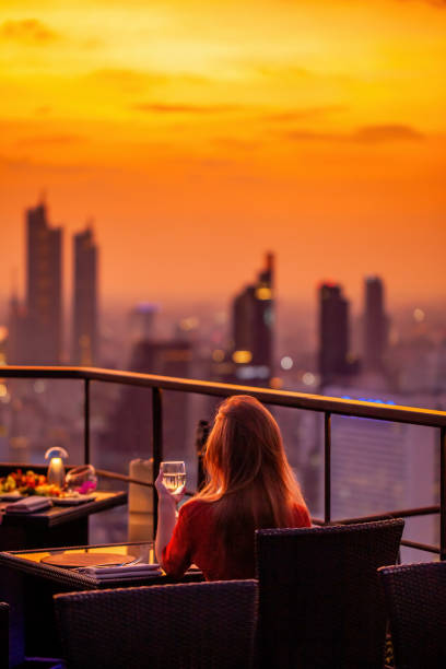 オレンジ色の空の夕日を眺める高級屋上レストランでワイングラスを飲んだ若い女性が休む。モダンな街のスカイラインを眺めるスカイバーテラスでカクテルドリンクを楽しめる女性。背景� - thailand restaurant cocktail bar ストックフォトと画像