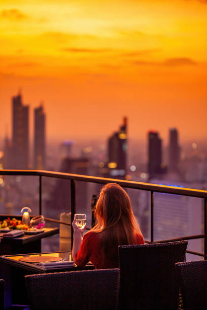 young woman with glass of wine rest at luxury rooftop restaurant watching orange sky sunset. female with cocktail drink at sky bar terrace looking at modern city skyline. skyscrapers on background - thailand restaurant cocktail bar imagens e fotografias de stock