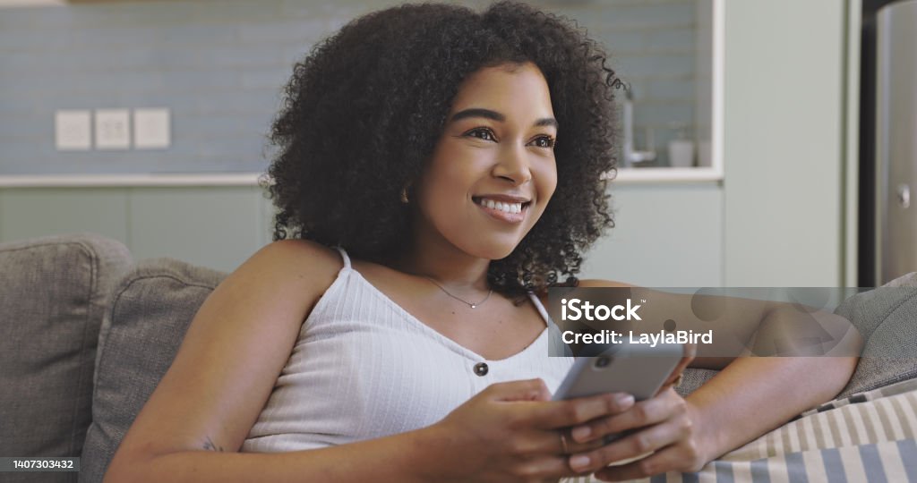 Beautiful young black woman using her phone while sitting on the couch in her living room at home. Happy female with afro thinking what to reply while texting or browsing through social media posts 20-24 Years Stock Photo