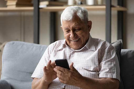 Happy elderly man look at cell phone screen rest on sofa at home, smiling enjoy received message with good news, read sms, share text messages spend leisure using modern wireless technology concept