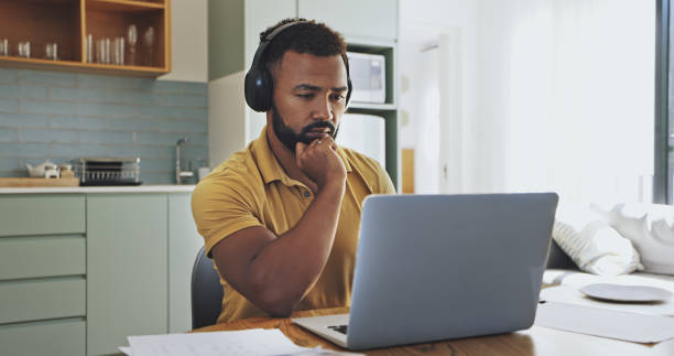 ein junger geschäftsmann, der seinen laptop benutzt und an etwas denkt, während er sitzt und lange stunden von zu hause aus arbeitet und auch ein headset in einer ernsten stimmung trägt und versucht, das problem zu lösen - laptop adult curtain business stock-fotos und bilder