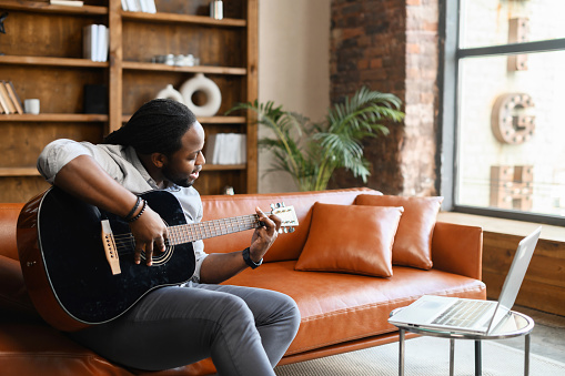 Concentrated young African American guy learning how to play guitar, watching online tutorial virtual class via laptop at home during coronavirus, sitting on the sofa, recording song streaming music