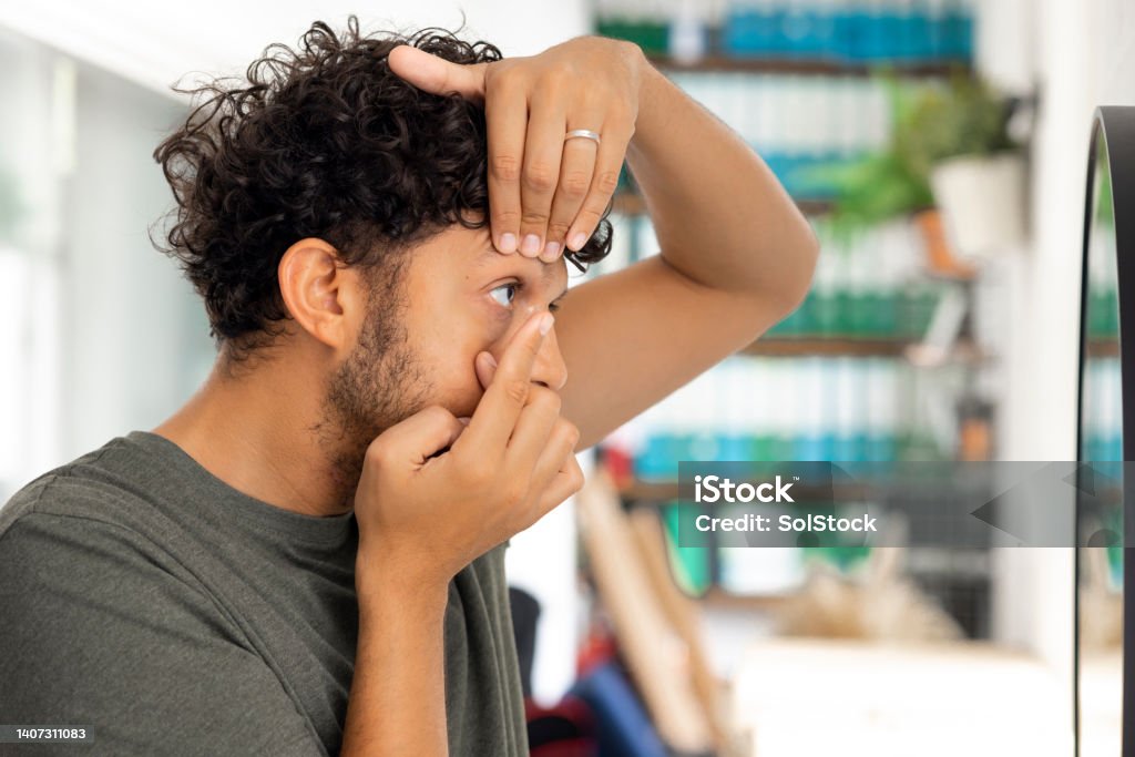 Contact Lens Application Side view medium shot a young male adult applying a contact lens looking in a vanity mirror wearing casual clothing. Contact Lens Stock Photo
