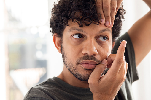 Close up of a young male adult applying a contact lens to his left eye wearing casual clothing.