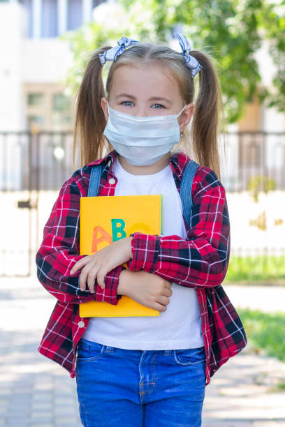 istock happy schoolgirl in a protective mask with a backpack and a textbook in her hands. in a white T-shirt and a plaid shirt 1407312664