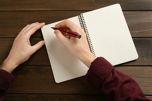 Woman writing with pen in notebook at wooden table, top view