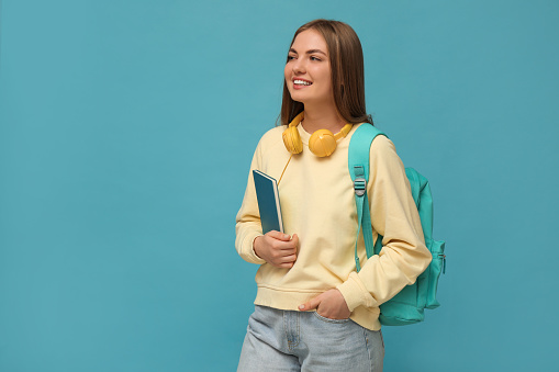 Teenage student with headphones, backpack and book on light blue background
