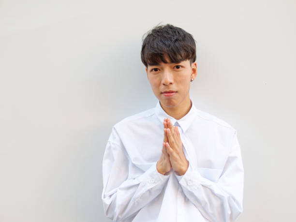 Portrait of Chinese young man with black hair in white shirt posing against white background, hands together prayer gesture, front view. stock photo
