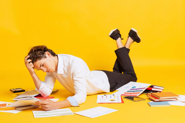 istock Young boy, student in business style clothes lying on floor and doing homework isolated over yellow background. Back to school, university 1407319531