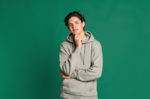 Looks calm. Studio shot of young boy, teen in casual style clothes looking at camera isolated over green studio background. Back to school, emotions, youth, fashion concept. Copy space for ad