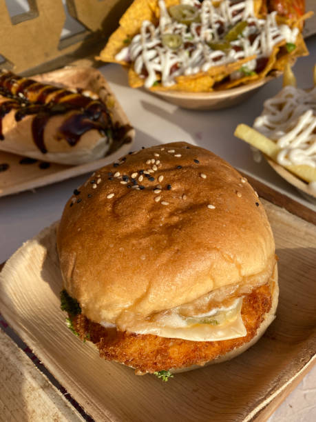 istock Close-up image of disposable bamboo plate containing chicken burger, sesame bread bun with breaded chicken breast, rocket, cheese slice, bowl of nachos, hotdog with barbecue sauce, french fries, restaurant setting, elevated view 1407322049
