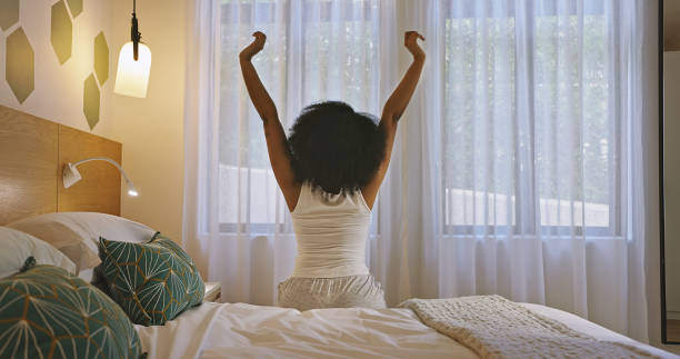 woman stretching arms to wake up and start a good morning on a bright new day from the back at home. one female with curly afro hair getting out of bed and feeling refreshed after some peaceful sleep - wake up stretching women black imagens e fotografias de stock