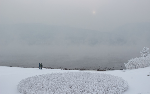 Early foggy winter morning at bank of the Yenisey river. Winter landscape with couple people among snow looking on river