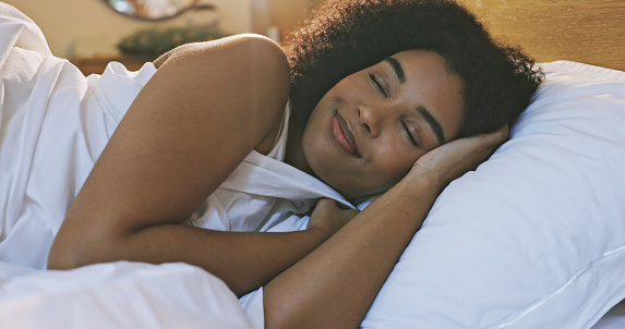 Woman with afro sleeping alone in her warm bed at home in the evening. Resting, dreaming, smiling in comfortable bedroom alone. Relaxed body, mind catching up on REM sleep for a healthy immune system