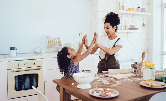 Little black playful joyful curly girl doing high five enthusiastically young cheerful happy mother during preparing cookies on large light kitchen at home