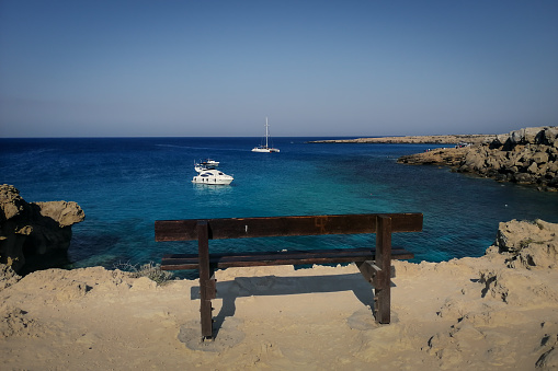 Empty wooden bench on the cliff with a view to the sea at Blue Lagoon, Cape Greco, Cyprus. Small boats anchored at the bay.