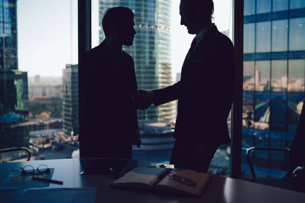 istock Formal businessmen standing in modern office at desk shaking hands 1407354563