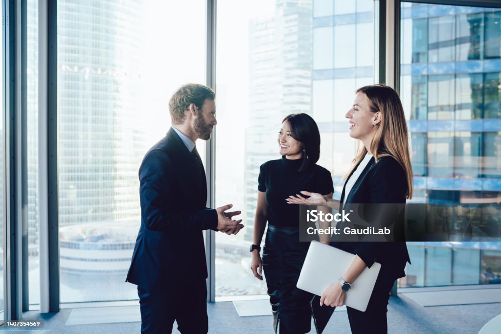 Young well dressed coworkers in office talking and laughing joyfully Diverse colleagues in elegant wear standing relaxed in circle in light big office sharing positive news and laughing with joy Corporate Business Stock Photo
