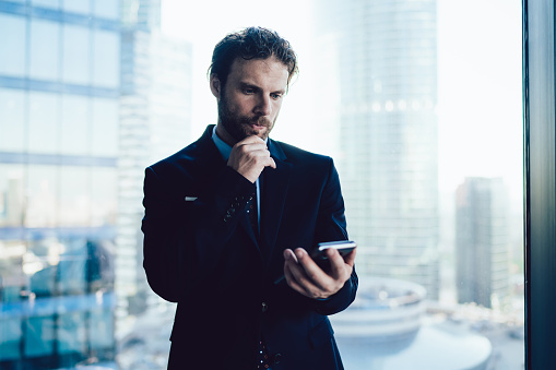 Formally dressed manager standing in front of window in office looking at cellphone with concentration while touching chin against blurred background