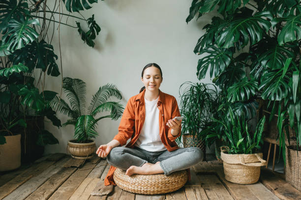 A beautiful young woman is sitting in meditation in the lotus position in a beautiful green garden among the plants of the house. The woman chooses music for meditation. The concept of mindfulness, psychological and mental health. stock photo