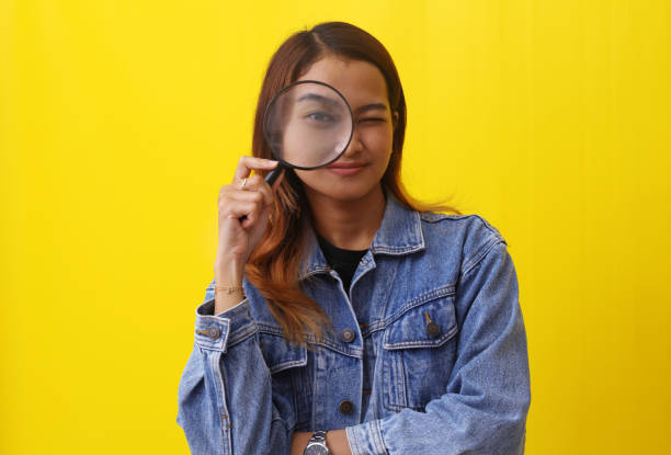 istock Cute asian woman standing while looking through magnifying glass. Isolated on yellow background 1407360409