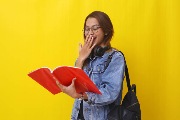 istock Amazed asian college student standing while open a book. Back to school concept. Isolated on yellow 1407361839