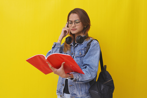 Thoughtful young asian college student standing while reading a book. Isolated on yellow background