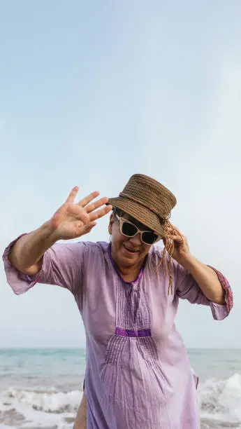 senior curvy woman in purple dress,sunglasses and hat waving while smiling happily on the beach in summer.vertical photo with copy space.travel and vacation concept.