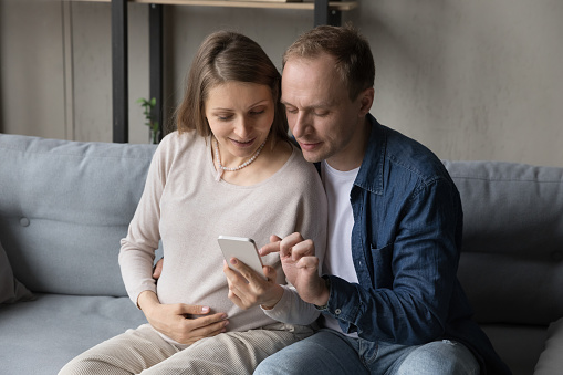 Young pregnant woman, her husband use smartphone sit on sofa at home. Mobile apps usage, pregnancy tracking, health-care tips. Family buy goods on internet, e-commerce, e-shopping, make order concept