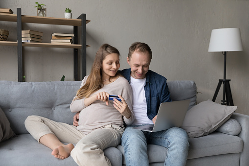 Pregnant wife her husband sit on sofa with laptop holds credit card buy goods for unborn baby on-line, make purchase, ordering on internet. Comfort life use modern tech, family distance e-shop concept