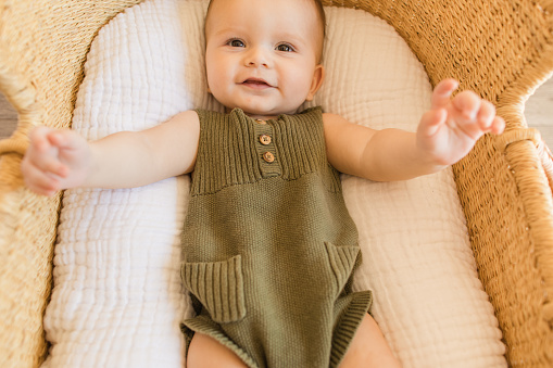 A Happy 27-Week-Old Baby Boy Smiling With His Feet Up in the Air, Wearing an Olive Green Knit Romper While Laying in a Cozy Cotton Blanket in a Seagrass Moses Basket in a Neutral Modern Home.