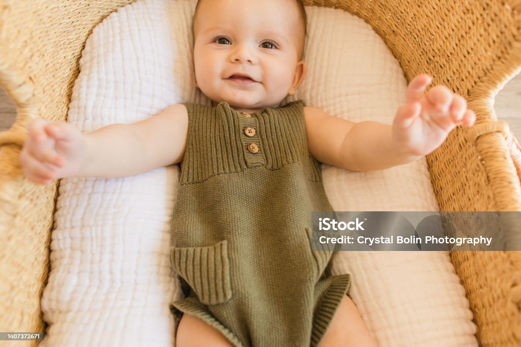 A Happy 27-Week-Old Baby Boy Smiling With His Feet Up in the Air, Wearing an Olive Green Knit Romper While Laying in a Cozy Cotton Blanket in a Seagrass Moses Basket in a Neutral Modern Home A Happy 27-Week-Old Baby Boy Smiling With His Feet Up in the Air, Wearing an Olive Green Knit Romper While Laying in a Cozy Cotton Blanket in a Seagrass Moses Basket in a Neutral Modern Home. Baby - Human Age Stock Photo