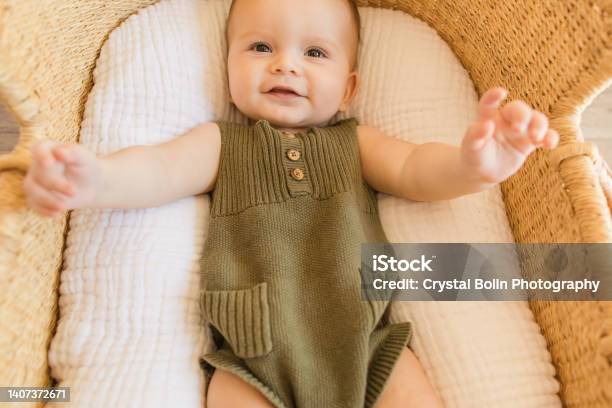 A Happy 27weekold Baby Boy Smiling With His Feet Up In The Air Wearing An Olive Green Knit Romper While Laying In A Cozy Cotton Blanket In A Seagrass Moses Basket In A Neutral Modern Home Stock Photo - Download Image Now
