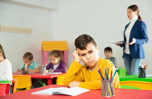 Portrait of upset boy in schoolroom on background with pupils studying with teacher